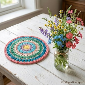 May include: A circular, crocheted doily in a vibrant palette of colours, including pink, blue, green, and yellow. A vase of wildflowers is next to it. The doily is on a white wooden table.