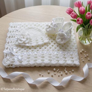 Puede incluir: Un conjunto de bebé blanco de ganchillo que incluye una manta, un gorro y patucos. La manta tiene cintas decorativas y detalles de flores. Los patucos y el gorro tienen adornos de perlas. Tulipanes rosas en un jarrón y perlas dispersas en el fondo.