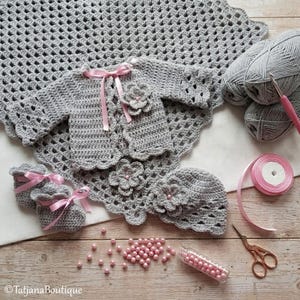 May include: A handmade crochet baby set in gray, including a blanket, jacket, booties, and hat, all adorned with pink ribbon and flower accents. The set is displayed with yarn, a crochet hook, and a spool of pink ribbon.