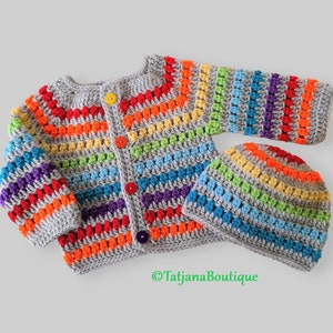 Op de afbeelding: Een grijs gebreid vest en bijpassende muts met regenboogstrepen. Het vest heeft knopen aan de voorkant.