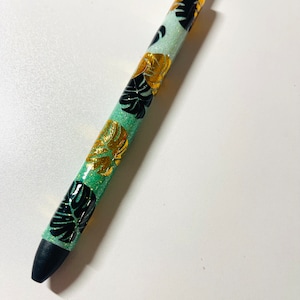 May include: A green pen with black and gold glitter and a tropical leaf design.