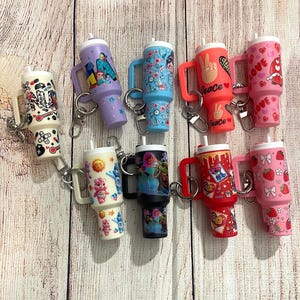 May include: Eight colorful mini tumblers with different designs, including cartoon characters, peace signs, and hearts. Each tumbler has a handle and a keychain attached.