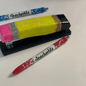 May include: A yellow, pink, silver and black glitter pencil-shaped stapler with a black base. Three pens with the text "#teacherlife" are also pictured.