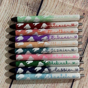 May include: Ten glitter pens with white tooth designs and personalized names, including "Tiana", "Martha", "Bihanna", "Jazmin", "Julian", "Shkinah", "Lassie", and "Alanta".