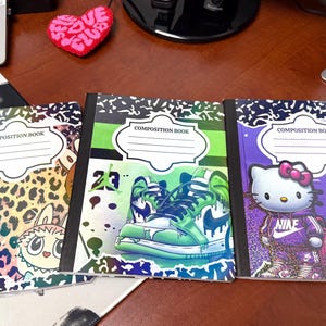 May include: Three composition notebooks with unique cover designs. One features cartoon characters and a leopard print background. Another showcases a green and blue sneaker design. The third notebook displays Hello Kitty in a Nike outfit.