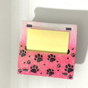 Può includere: Porta note da scrivania rosa e bianco glitter con design a forma di impronta di zampa nera. Un post-it giallo è nel porta note.