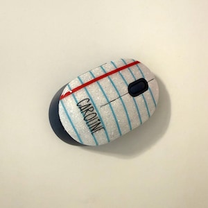 Può includere: Un mouse per computer wireless con finitura glitterata bianca, caratterizzato da un motivo a foglio di quaderno con linee blu orizzontali, una linea di margine rossa verticale e il nome CAROLINE stampato in nero. Include una rotellina blu scuro.