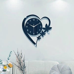 Peut inclure: Une horloge murale noire en forme de cœur avec un cadran circulaire et des chiffres blancs. L'horloge présente des accents floraux décoratifs et est fixée sur un mur blanc. Les aiguilles indiquent l'heure.