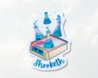 May include: A die-cut sticker featuring a laboratory hot plate with three glass Erlenmeyer flasks containing blue and green liquids. The flasks are floating above the hot plate, surrounded by small yellow stars. The text Shooketh is printed below.