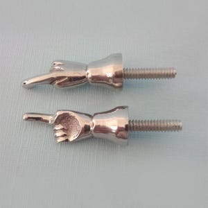 May include: Two silver-colored cabinet knobs shaped like hands pointing. Each knob has a threaded bolt for attachment. The knobs are on a light blue background. These unique knobs add a playful touch to furniture.