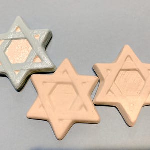 May include: Three Star of David ornaments in a light color palette. One is a muted gray, and the other two are a light peach color. Each has a six-pointed star shape with a geometric design in the center.