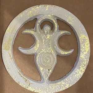 Puede incluir: Un colgante de pared circular blanco con un acabado de hoja de oro. El diseño presenta una figura femenina estilizada con tres lunas crecientes y una espiral.