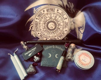 May include: A collection of items including a circular wooden calendar, two light gray candles, a black taper candle, a small black tin labeled Hedge Riding Ointment, a square slate coaster with a key, a roller bottle, and a round tin.