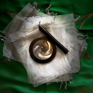 May include: A black circular mirror with a swirling galaxy design inside, a black candle, and a stack of white organza bags.