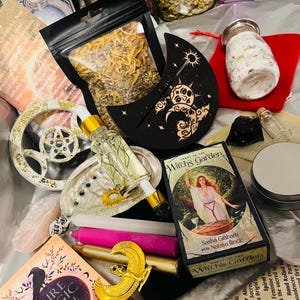 May include: A collection of witchy items including a deck of tarot cards, a moon-shaped wooden sign, a bag of dried herbs, a bottle of essential oil, a candle, a crystal, and a small jar of bath salts. The tarot cards are from the "Tarot of the Witch's Garden" deck. The moon sign has a sun and stars design. The bag of herbs is labeled "Chamomile".