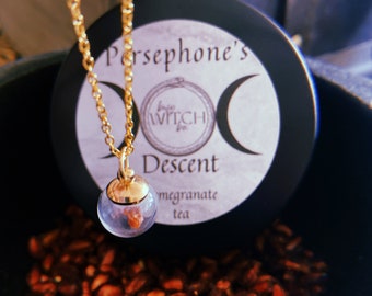 May include: A gold chain necklace with a clear glass pendant containing a small piece of pomegranate. The necklace is resting on a black container with a white label that reads "Persephone's  Into Witch  Descent  Pomegranate Tea".