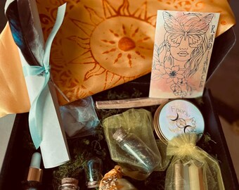 May include: A gift box containing a yellow sun-patterned cloth, a scroll with a dark feather tied by a light blue ribbon, a card with a butterfly-faced illustration labeled Radiance, a gold bell, glass vials with contents, and candles in a yellow bag.