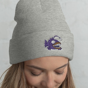 Puede incluir: Un gorro gris con un gráfico de pez pescador morado. El pez pescador tiene un señuelo blanco y una sonrisa de dientes.