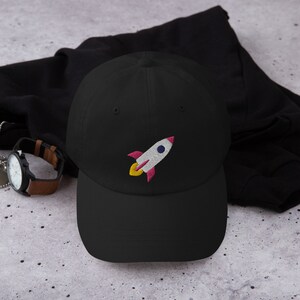 May include: Black baseball cap with an embroidered rocket ship design in white, pink, and yellow.