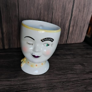 May include: A white ceramic face-shaped cup with a yellow rim and a yellow scarf with orange polka dots. The cup has a winking eye, a closed eye, and the word "Baileys" printed on the front. The face has pink cheeks and a purple lip.