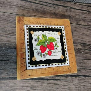 May include: Square wooden coaster with a decorative tile insert. The tile features a black background with a white, scalloped edge, and a vibrant illustration of red strawberries, green leaves, and white flowers. Handwritten script surrounds the image.