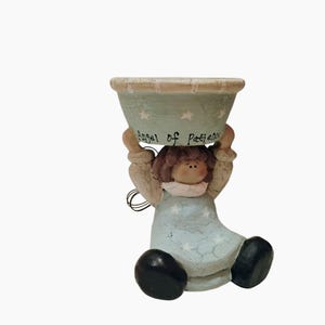 Może przedstawiać: Ceramiczna figurka anioła trzymającego misę nad głową. Misa jest jasnoniebieska z białymi gwiazdami i napisem "Angel of Patience". Anioł ma na sobie jasnoniebieską sukienkę i czarne buty.