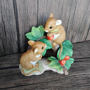 May include: A ceramic figurine featuring two brown mice on a branch with green leaves and red berries. One mouse holds a berry. The figurine is painted with detailed features and sits on a light-colored base. The background is a wooden surface.