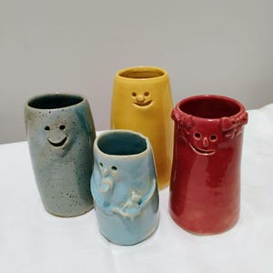 Peut inclure: Quatre récipients en céramique de forme cylindrique avec des visages sculptés. L'un est gris moucheté, un jaune vif, un bleu clair avec une petite figurine attachée, et un rouge avec des nœuds décoratifs sur les côtés du rebord.