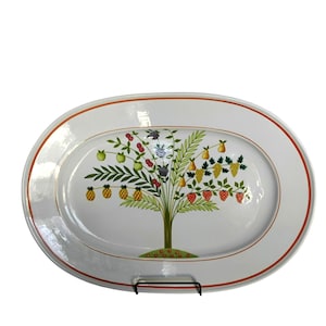 May include: Oval white serving platter with a tree design featuring various fruits. The tree has a green trunk and branches, with pineapples, apples, pears, grapes, and strawberries. The platter has a red-orange border and is supported by a black metal stand.