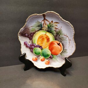 May include: A decorative porcelain plate featuring a still life of fruit. The plate has a scalloped edge with gold trim. The painted image includes a cantaloupe, peach, grapes, and cherries. The plate is displayed on a black stand.