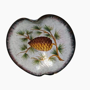 May include: A heart-shaped decorative dish with a white speckled interior. The dish features a painted design of a pine cone on a branch with green needles. The rim is dark brown with a lighter brown edge.