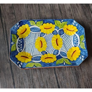 May include: A rectangular ceramic tray featuring a hand-painted pattern of bright yellow sunflowers with brown centers, green leaves, and blue accents. The background displays a blue and white cross-hatched grid design with decorative blue borders.
