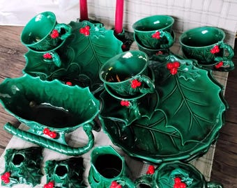 Può includere: Un set di stoviglie in ceramica verde con motivi a foglie di agrifoglio in rilievo e bacche rosse brillanti. La collezione include tazze, piattini, una grande ciotola da portata, una lattiera, una zuccheriera con coperchio e due candelabri.