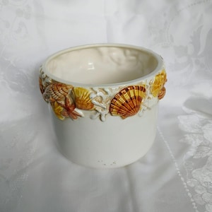 May include: A cylindrical ceramic planter with a smooth off-white finish. The upper rim is decorated with a raised, three-dimensional relief of various seashells and coral in shades of golden yellow, orange, and brown, encircling the top edge.