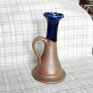 Puede incluir: Un candelabro de cerámica con una base ancha y acampanada y un cuello alto y esbelto. La parte inferior tiene un acabado marrón terroso y texturizado, mientras que el cuello superior y el borde están esmaltados en un azul cobalto brillante.
