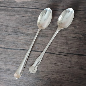 May include: Two silver-toned metal spoons with long, slender handles. The spoon on the right features an ornate, embossed shell-like pattern at the base of the handle, while the spoon on the left has a smooth, plain handle design.