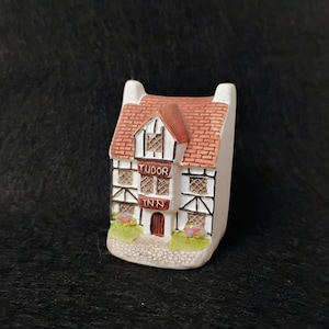 Puede incluir: Una figura en miniatura de la posada Tudor. El edificio presenta un tejado de tejas rojas, paredes blancas con entramado de madera marrón y una pequeña puerta. El texto "TUDOR INN" se muestra en la parte delantera. Acentos florales verdes y morados en la base.