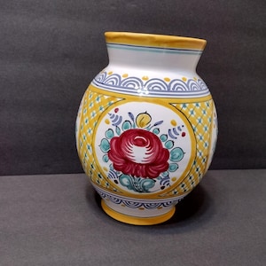 May include: A ceramic vase featuring a hand-painted design with a central red rose motif surrounded by teal leaves and yellow accents. The body displays a yellow and white checkered pattern, bordered by blue scalloped lines and a yellow rim.