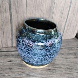 Pode incluir: Um pequeno vaso de cerâmica redondo com um esmalte azul e lavanda salpicado. O vaso tem um interior escuro e uma base de cor clara. A borda é um tom mais escuro de azul. O vaso está sobre uma superfície de madeira.