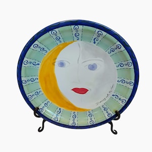 May include: A decorative ceramic plate featuring a celestial design. The plate has a light green background with a blue border and a stylized face of the moon. The moon has a yellow crescent, white face, red lips, and blue eyes. The text "Dancing with the Moon" is written on the plate.