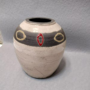 Könnte beinhalten: Eine handgefertigte Keramikvase mit cremefarbenem Körper, einem dunkelgrauen Band und einem schwarzen Innenraum. Die Vase hat ein rotes Oval und zwei gelb umrandete Kreise. Die Vase ist etwa 15 cm hoch.