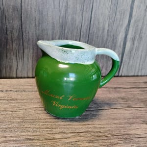 Puede incluir: Jarra pequeña de cerámica verde con borde y asa blancos. Las palabras "Mount Vernon Virginia" están escritas en letras doradas en la parte delantera. Probablemente un recuerdo o un artículo decorativo.