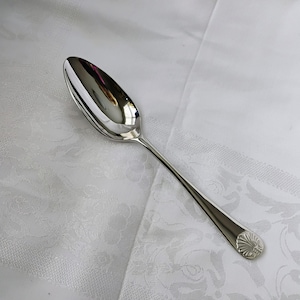 Może przedstawiać: Polerowana srebrna łyżka do serwowania z długim trzonkiem i wzorem muszli na końcu. Łyżka ma dużą, owalną miseczkę i odbija otaczające światło. Łyżka leży na białym obrusie.