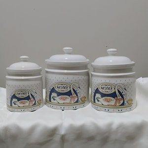 May include: A set of three white ceramic canisters with lids, featuring a repeating pattern of small blue hearts and a central illustration of a white swan wearing a pink bow, with the word HOME printed above the swan in a decorative oval frame.