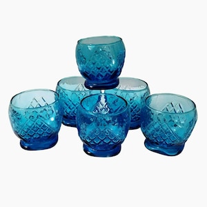 Puede incluir: Seis portavelas votivas de cristal azul. Cada uno tiene un patrón de diamantes y una forma redondeada. Los portavelas están dispuestos en grupo, con uno apilado encima de los demás.