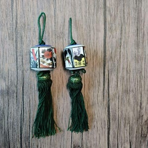 May include: Two decorative hanging ornaments featuring cylindrical ceramic beads with floral and bird illustrations, black text, and green cord loops. Each bead is topped with a green cord loop and finished with a long, dense green tassel.