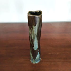 May include: A tall, slender ceramic vase with an irregular, organic shape. The surface features a reactive glaze with deep brown, dark chocolate tones dripping over a muted, dusty blue-green base, creating a layered, textured appearance.
