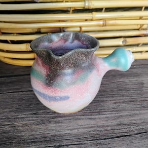 May include: A small ceramic creamer or jug with a rounded body and a single side handle. The exterior features a mottled glaze with blended hues of dark brown, muted green, soft pink, and light blue, with a wavy, uneven rim.