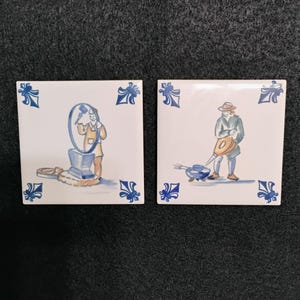May include: Two square ceramic tiles with white backgrounds and blue and white illustrations. One tile depicts a person working with a machine, the other shows a person with a watering can. Each tile has blue floral corner designs.