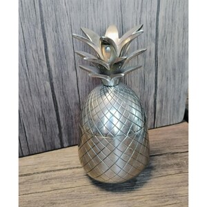 Puede incluir: Un recipiente plateado con forma de piña y superficie texturizada. El recipiente tiene tapa y un diseño detallado, que se asemeja a una piña real. El recipiente es de metal y está sobre una superficie de madera.